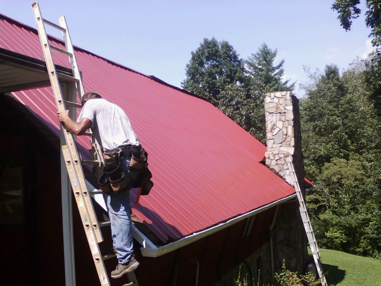Roofer on a ladder beside a red metal roof and stone chimney — Soffit & Fascia Repair by Roofing Brothers in Leeds